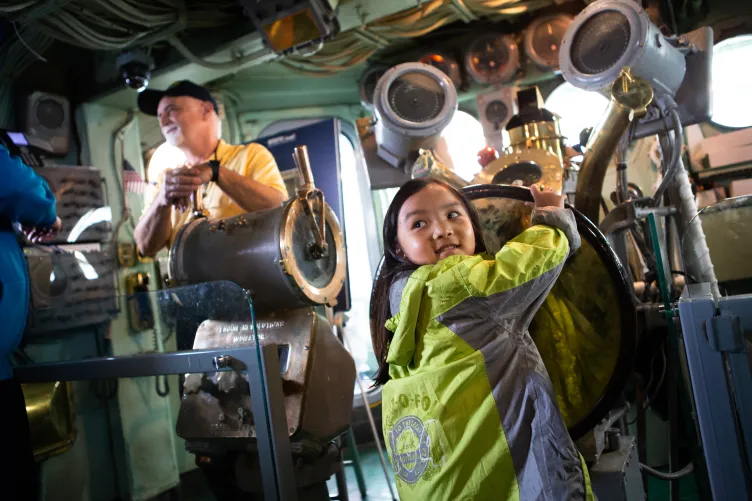 A young girl plays in the ship's island, spinning the ship's wheel as a former Intrepid crew member looks on