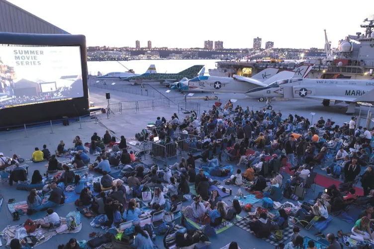 A large group of people are watching a movie on the flight deck of an aircraft carrier, surrounded by planes. 