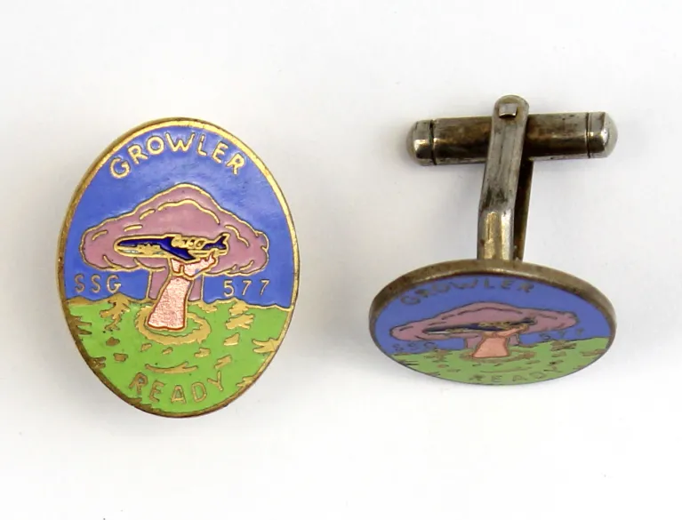 The front and sides of an old pin that has an arm outstretched holding a blue submarine.