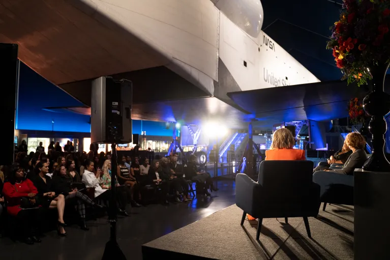 A diverse group of people seated beneath the Space Shuttle pavilion, gathered for a discussion.