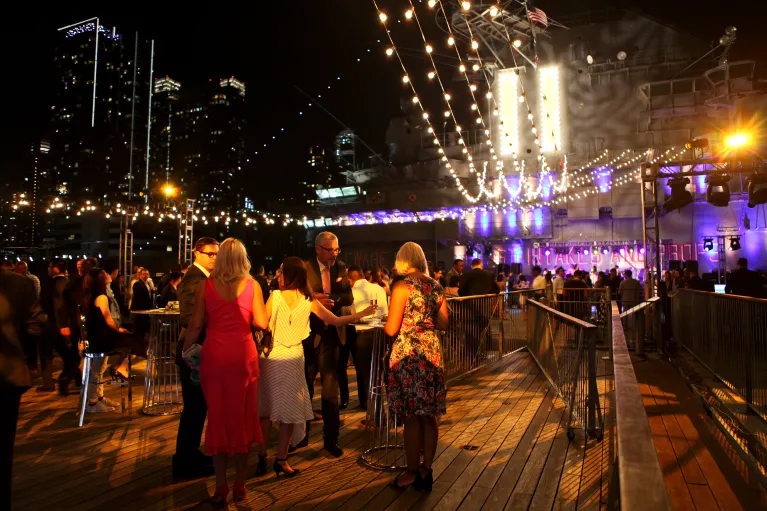 Guests gather for a cocktail reception on the port side aircraft elevator at flight deck level with string lighting above and views of the city skyline