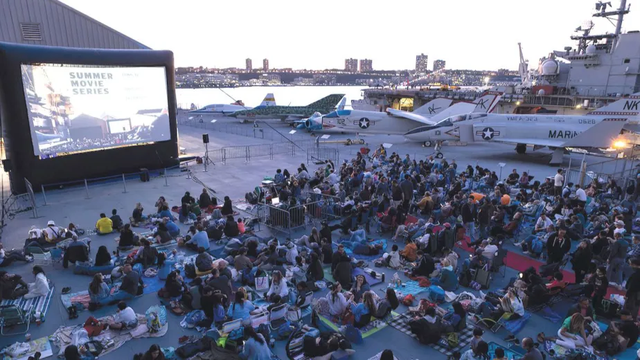 A large group of people are watching a movie on the flight deck of an aircraft carrier, surrounded by planes.