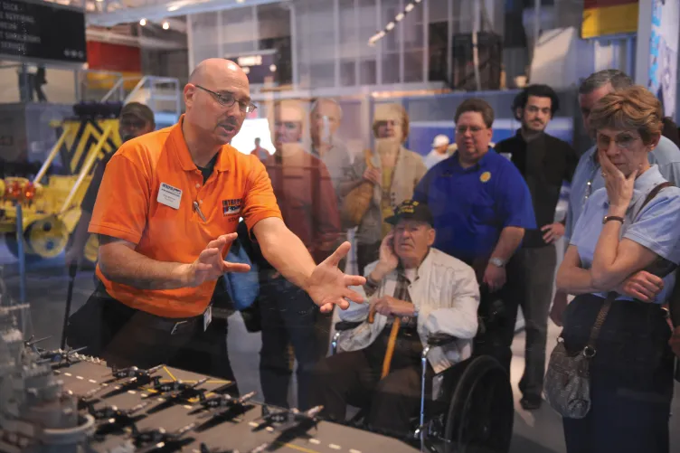 A tour guide is speaking to a group of visitors in front of a model of Intrepid.