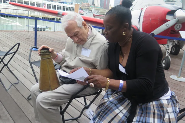 A woman is helping an older man read something while he holds a large horn.