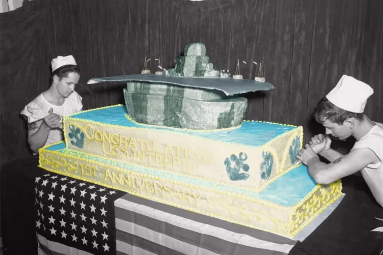 Archival photo of two people decorating a very large, double layer cake with an aircraft carrier on top.