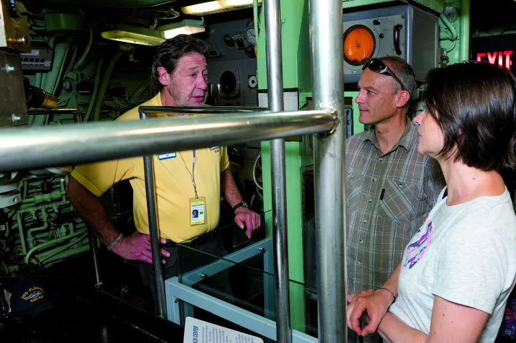 A veteran is speaking with two visitors next to the Growler's periscope