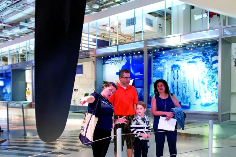 A tour guided is speaking with several visitors next to a propeller