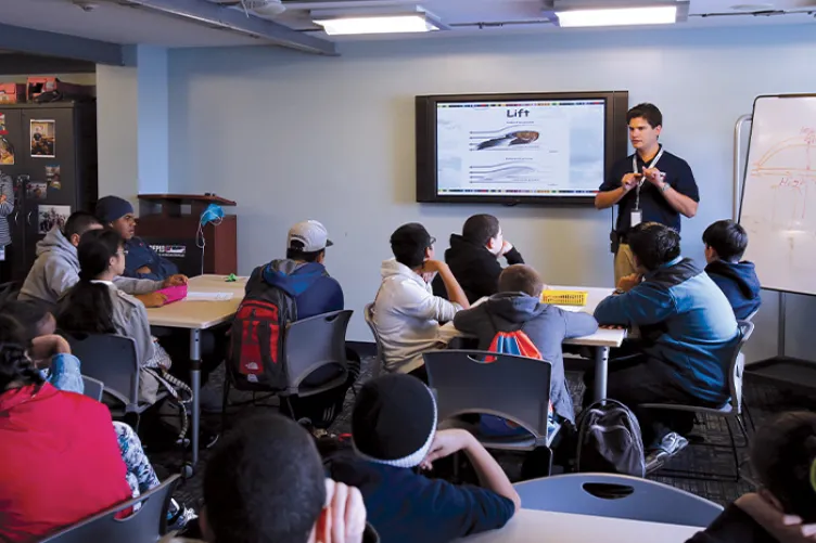 An educator is in a classroom in front of a screen speaking to a group of students.