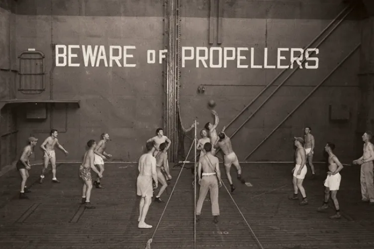 Archival photo of men playing volleyball on the flight deck with words painted behind them that say "Beware of Propellers"