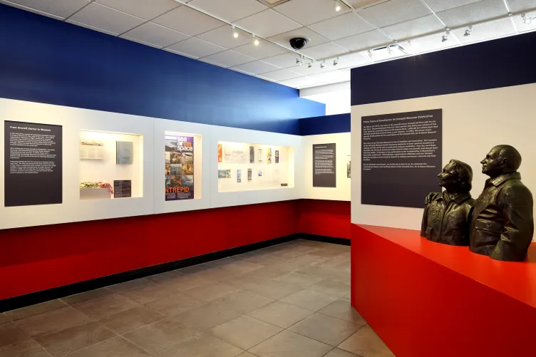 Sculptures of two people smiling and looking outward towards the exhibition panels, highlighting the Intrepid Museum’s history.