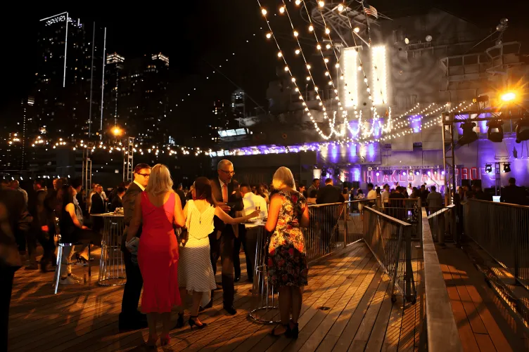 People gather on the Port Side Aircraft Elevator during a night time cocktail reception, with drinks and string lights above from the Flight Deck Island