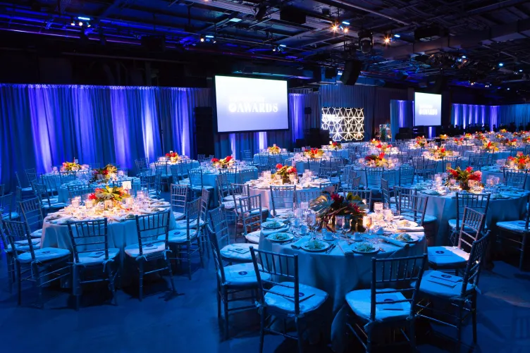 Hangar 3 setup for a gala dinner and program with blue lighting, red florals on dinner tables, and audio-visual screens at the side of the stage for the program.