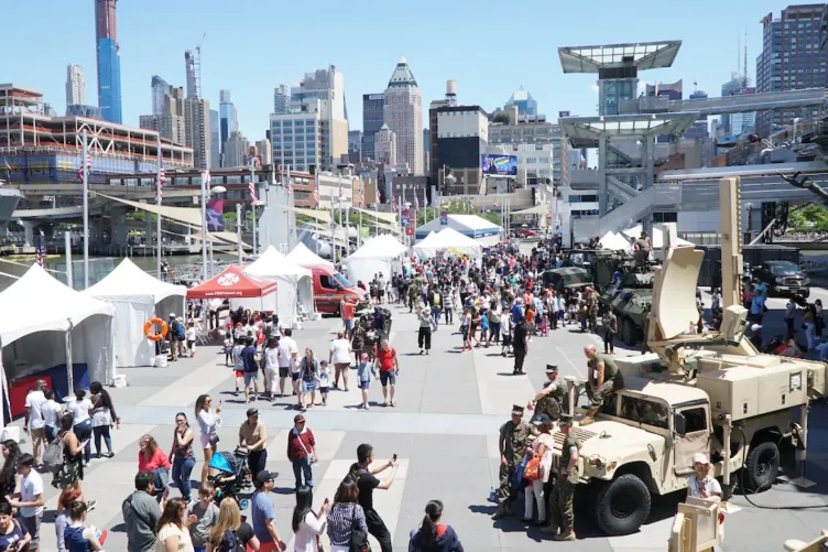 Crowds of people on Pier 86 during Fleet Week, with white event tents on the left, and active duty military personnel are sitting on military vehicles on the right.