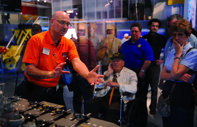 A Museum educator is standing behind a table that has a globe and a model rocket on it and is speaking to a group of seated visitors.