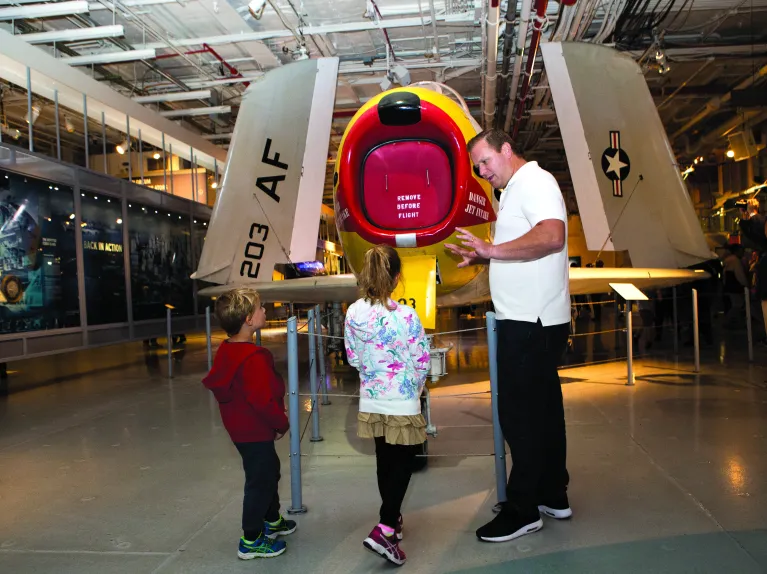 An adult speaking to two children in front of an airplane.