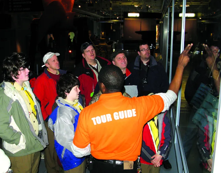 A tour guide is speaking to a group of visitors in front of an exhibition panel.