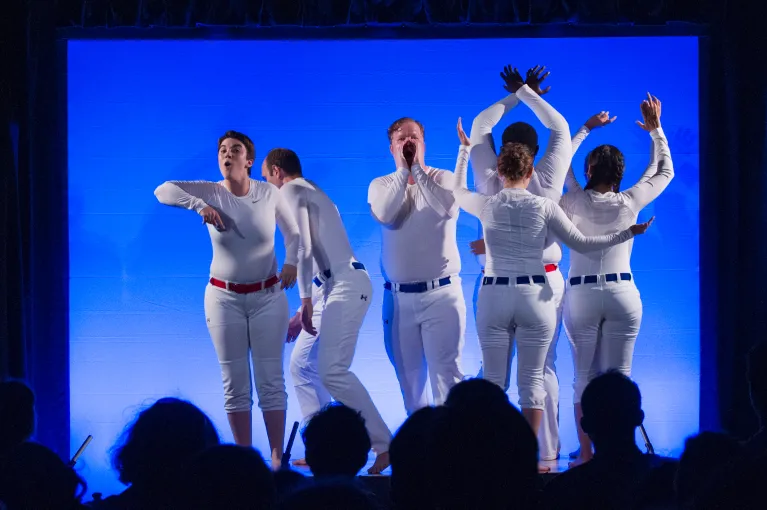 Performers dressed on white with a blue background on stage perform for a group of visitors.
