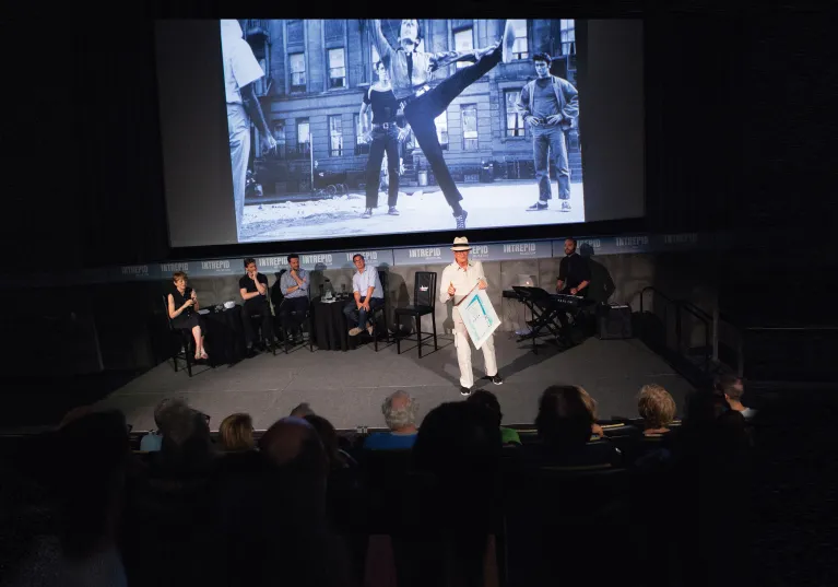 A group of visitors at the Allison & Howard Lutnick Theater listen to a group of panelists on the stage with a large screen in the background.