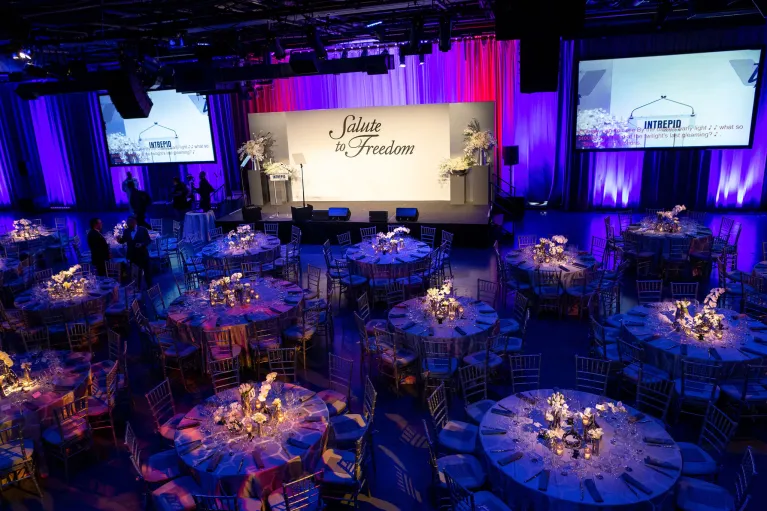 Hangar 3 setup for a gala dinner and program with a stage, dinner tables, AV screens, uplighting, and florals