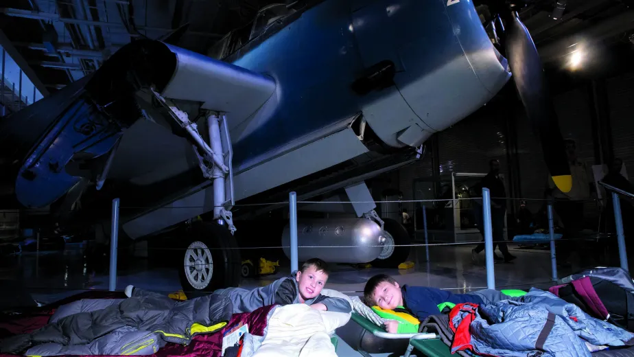 Two kids in sleeping bags are laying on top of cots on the hangar deck next to an airplane