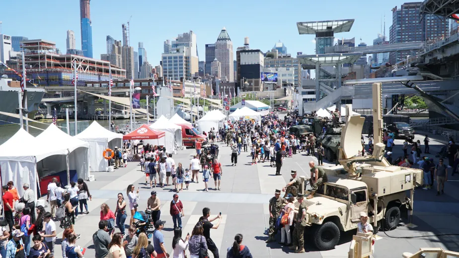 Crowds of people are on Pier 86, with white event tents on the left, and active duty military personnel are sitting on military vehicles on the right.