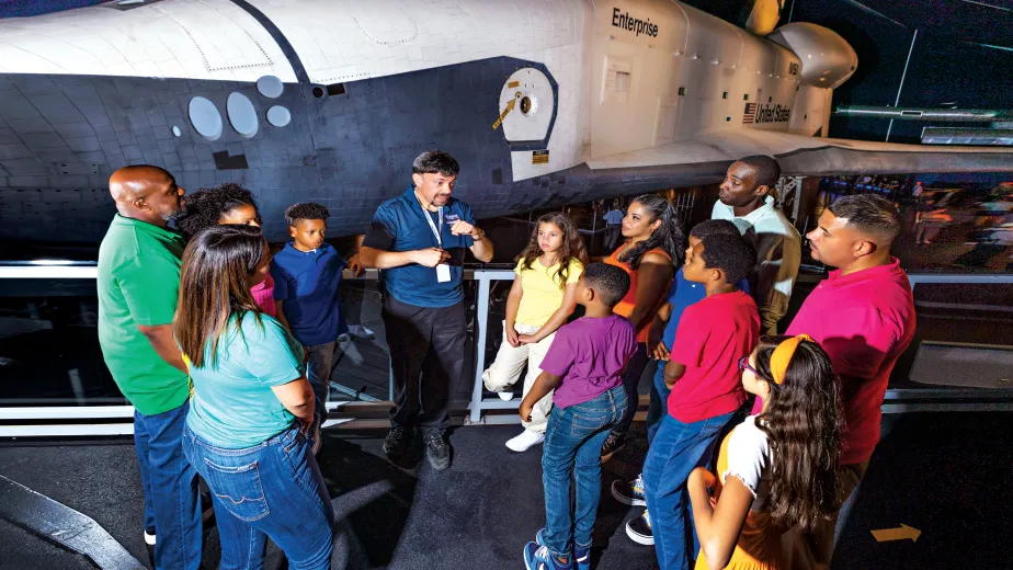 A group of visitors stand in front of the Space Shuttle Enterprise and are listening to a tour guide.