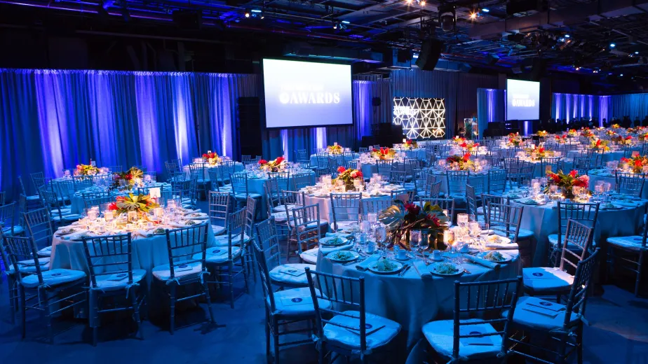 Hangar 3 setup for a gala dinner and program with blue lighting, red florals on dinner tables, and audio-visual screens at the side of the stage for the program.